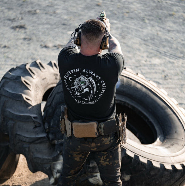 Person with headphones and a tactical gear standing between two large tires on a gun range.