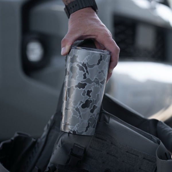 Hand holding a Savage Tacticians camo tumbler next to tactical gear outdoors