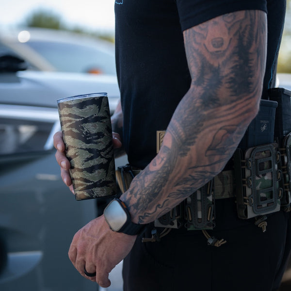 Tattooed man holding Savage Tacticians camo tumbler, tactical gear visible outdoors