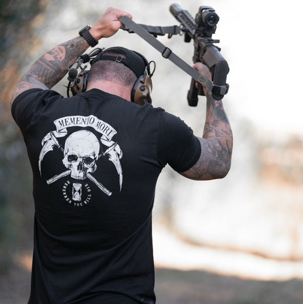 Man holding a rifle with a black t-shirt featuring a skull and scythe design on the back. Memento Mori.