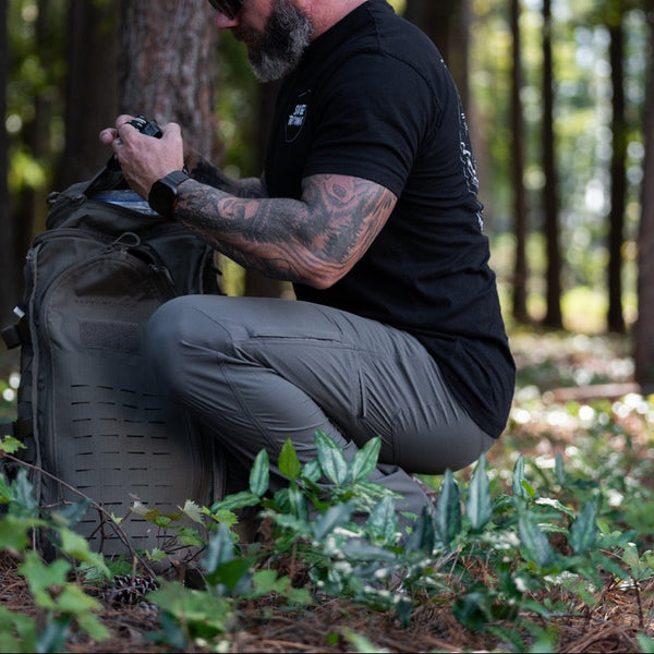 Savage Tacticians shirt worn by bearded man with tattoos kneeling in forest with backpack