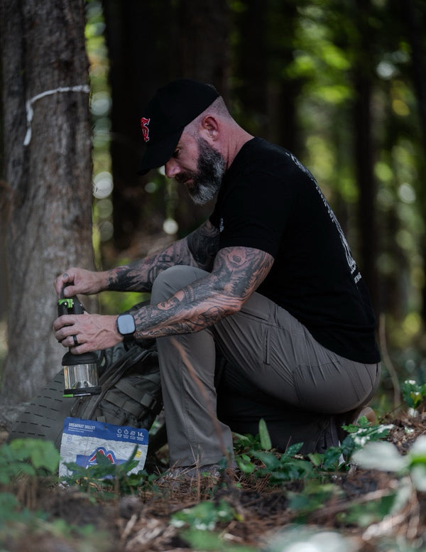 Tattooed man in Savage Tacticians apparel outdoors with lantern and camping gear
