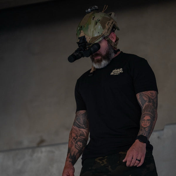 Tattooed man in Savage Tacticians shirt, camo helmet, night vision goggles indoors