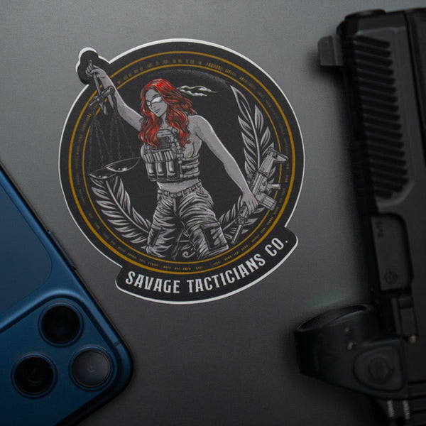 Savage Tacticians sticker featuring armed woman with scales of justice, on laptop near phone and gun
