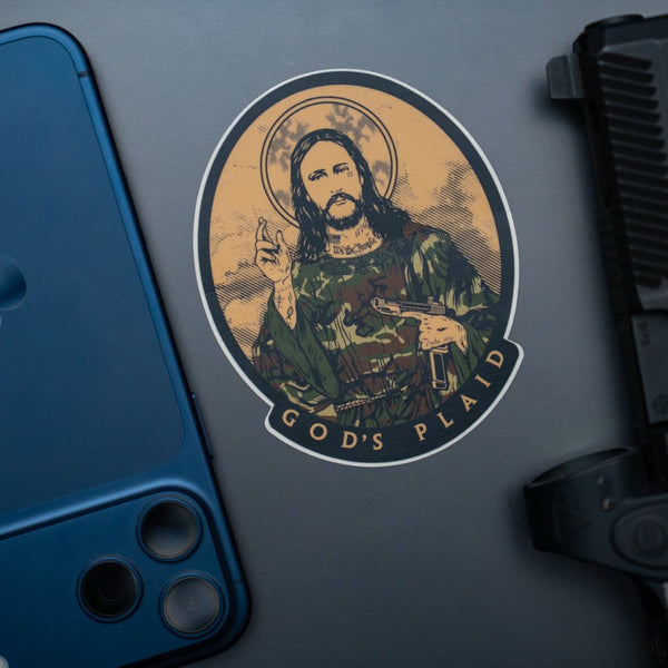 Savage Tacticians sticker of a saint in camo with 'God's Plaid' text, next to phone and pistol