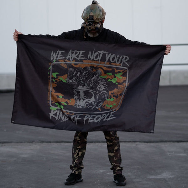 Person in tactical gear holding Savage Tacticians flag with skull graphic and camo design