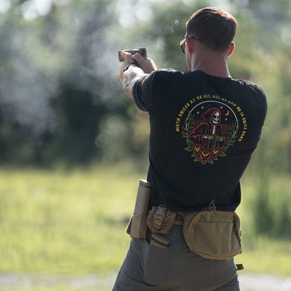 Man at outdoor range wearing Savage Tacticians graphic tee, shooting pistol, tactical gear visible