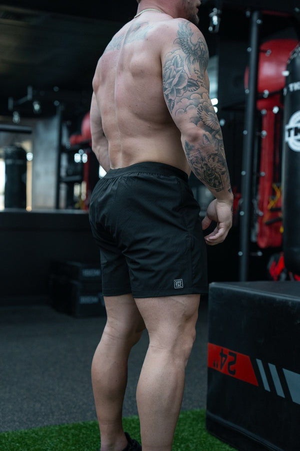 Muscular man with tattoos wearing Savage Tacticians black athletic shorts in gym