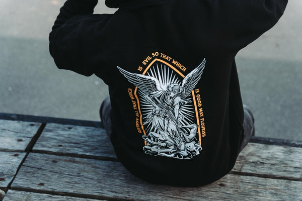 Man seated on a wooden deck showing the archangel back print of the black Savage Tacticians Saint Hoodie