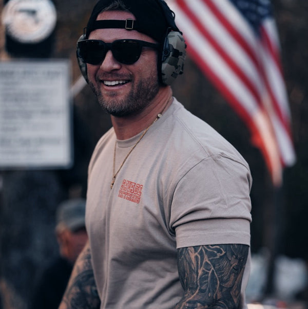 Man wearing Savage Tacticians t-shirt, sunglasses, and headset outdoors with American flag