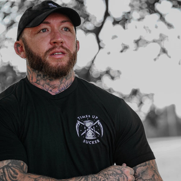 Man wearing a black t-shirt with a Savage Tacticians graphic tee, standing against a blurred abstract background.