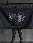 Person in camo pants holding Savage Tacticians flag with skeleton art and urban background