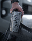 Hand holding a Savage Tacticians camo tumbler next to tactical gear outdoors