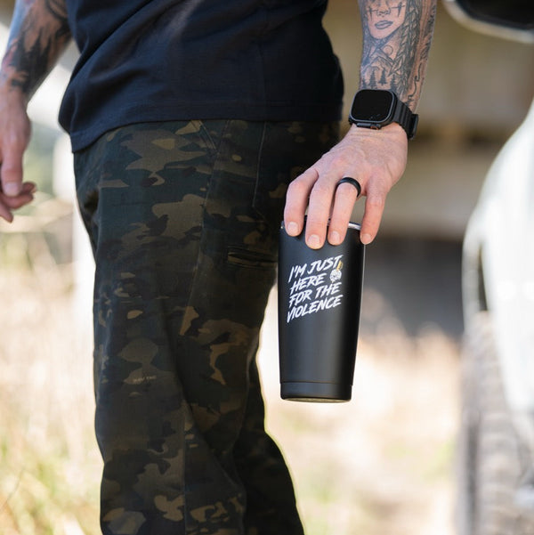 Man with tattooed arm holding Savage Tacticians tumbler, camo pants, outdoor setting