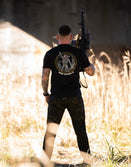 Man outdoors holding rifle, wearing Savage Tacticians black graphic t-shirt and camo pants