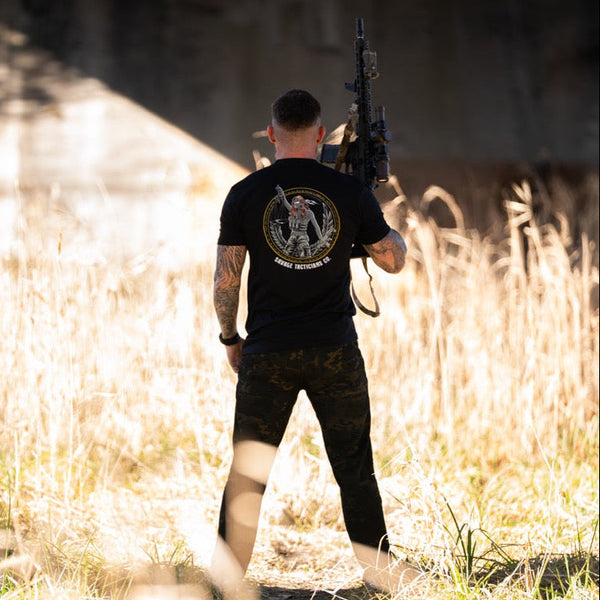 Man outdoors holding rifle, wearing Savage Tacticians black graphic t-shirt and camo pants