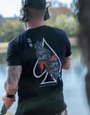 Man wearing Savage Tacticians t-shirt with ace of spades and skull graphic, outdoors by water