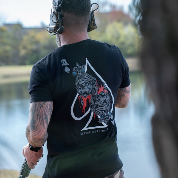 Man wearing Savage Tacticians t-shirt with ace of spades and skull graphic, outdoors by water