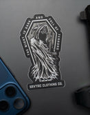 Savage Tacticians sticker with grim reaper design and text, on dark surface near phone