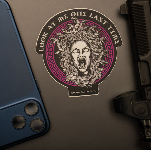 Savage Tacticians Medusa sticker on laptop, beside blue smartphone and gun