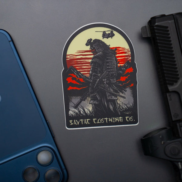 Savage Tacticians sticker of a modern samurai with gear, sunset background, phone and pistol nearby