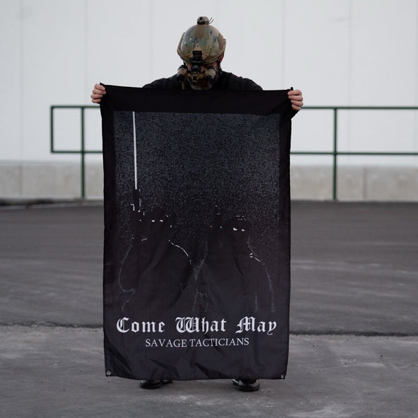 Person in tactical helmet holding Savage Tacticians 'Come What May' black flag outdoors