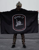 Person in camo pants and tactical helmet holding Savage Tacticians SEND IT flag outdoors