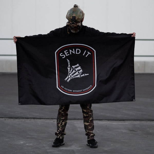 Person in camo pants and tactical helmet holding Savage Tacticians SEND IT flag outdoors