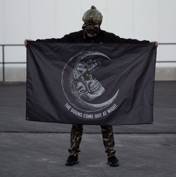 Person in tactical helmet holding Savage Tacticians flag with skull moon and 'The Goons Come Out At Night' text