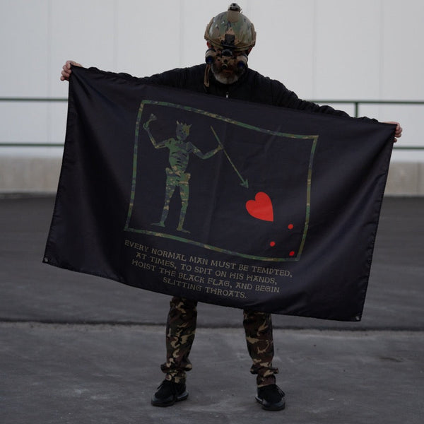 Person in tactical gear holding Savage Tacticians flag with skeleton and heart graphic.