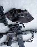 Savage Tacticians camouflage boonie hat with SavTac logo beside tactical firearms on concrete