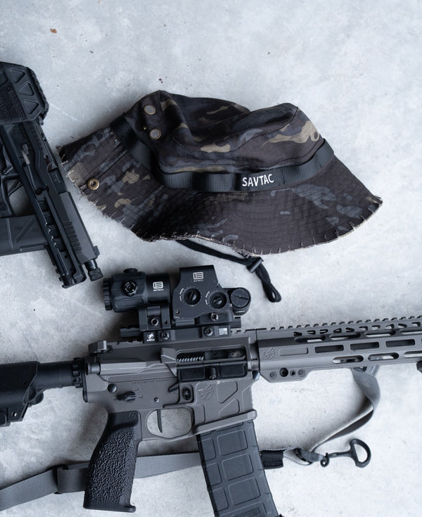 Savage Tacticians camouflage boonie hat with SavTac logo beside tactical firearms on concrete