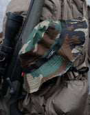 Savage Tacticians camo bucket hat with black logo on a tactical backpack and rifle