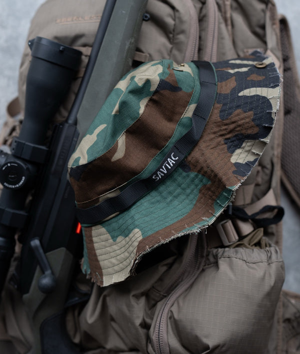 Savage Tacticians camo bucket hat with black logo on a tactical backpack and rifle