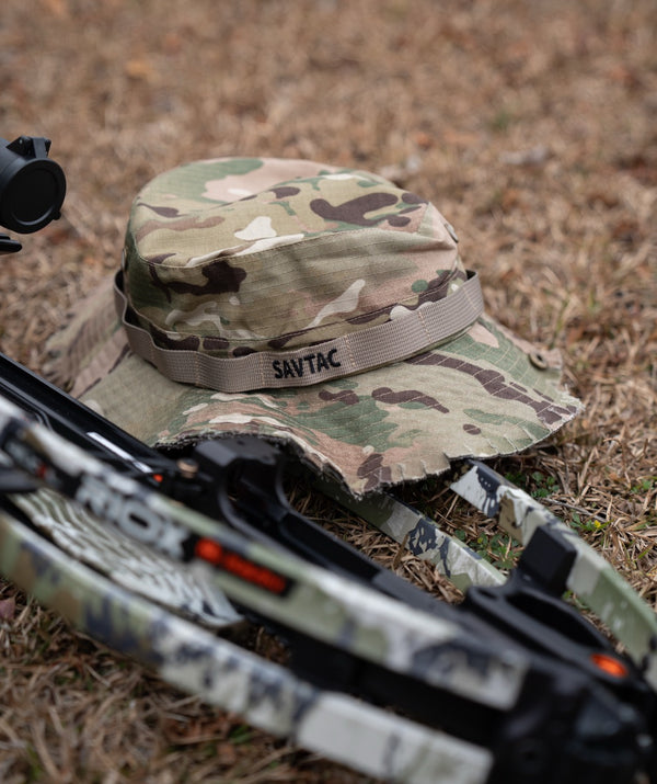 Savage Tacticians camo boonie hat on grass next to a compound bow, outdoor setting