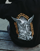 Man seated on a wooden deck showing the archangel back print of the black Savage Tacticians Saint Hoodie