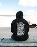 Man seen from behind sitting outdoors in the black Savage Tacticians Saint Hoodie