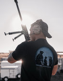 Man outdoors in headset and black Savage Tacticians t-shirt holding rifle, sunlight behind