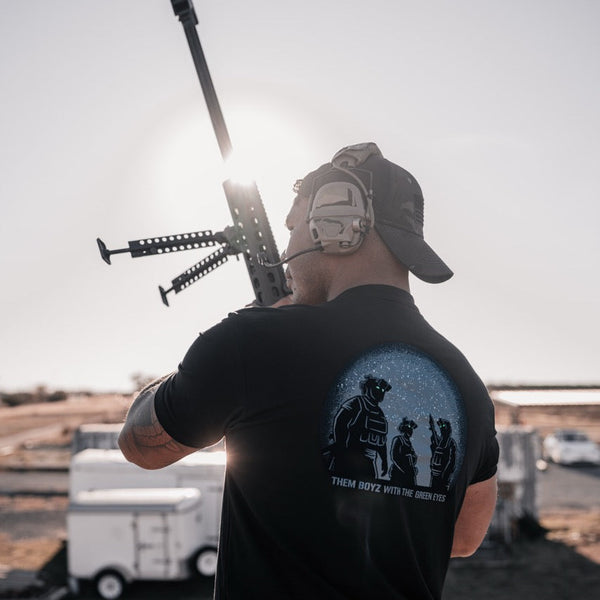 Man outdoors in headset and black Savage Tacticians t-shirt holding rifle, sunlight behind