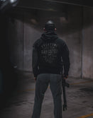 Man seen from behind in a dark garage showing the lettered back print of the black Savage Tacticians Gangster Hoodie