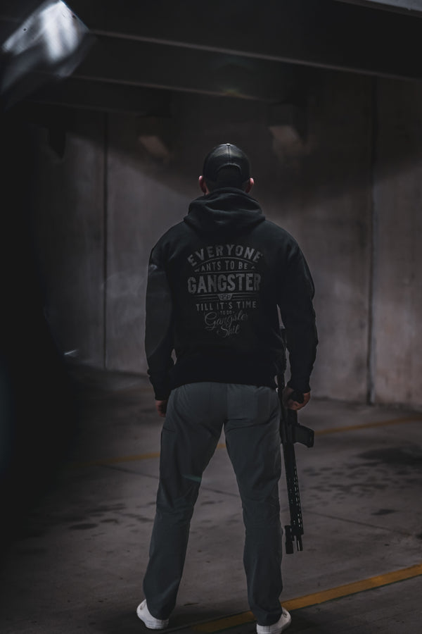 Man seen from behind in a dark garage showing the lettered back print of the black Savage Tacticians Gangster Hoodie