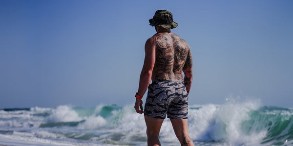 Savage Tacticians swim trunks camo on tattooed man at the beach with ocean waves